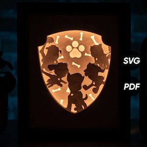 May include: A black and white silhouette of the Paw Patrol characters, including Chase, Marshall, Skye, Rubble, Rocky, and Zuma, arranged in a circle around a paw print. The characters are inside a shield shape with a white background and bone-shaped cutouts. The image is labeled "SVG" and "PDF" in the bottom right corner.