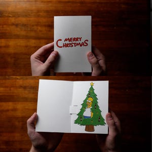 May include: A white Christmas card with the words "MERRY CHRISTMAS" in red. The inside of the card features a cartoon image of Homer Simpson as a Christmas tree, decorated with beer bottles. The card is held open, revealing the illustration.