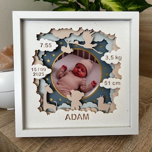 Op de afbeelding: Een witte schaduwdoos met een babyfoto, omringd door uitgesneden vormen van wolken, dieren en een ooievaar. De lijst bevat de geboortegegevens van de baby: 7:55, 15/09/2025, 3,5 kg en 51 cm. De naam van de baby, ADAM, staat onderaan.