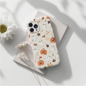 May include: A phone case with a Halloween theme. The case has a cream background with a pattern of cartoon ghosts, orange pumpkins, and brown leaves. The phone case is on a white surface with a white flower and a sprig of white flowers.