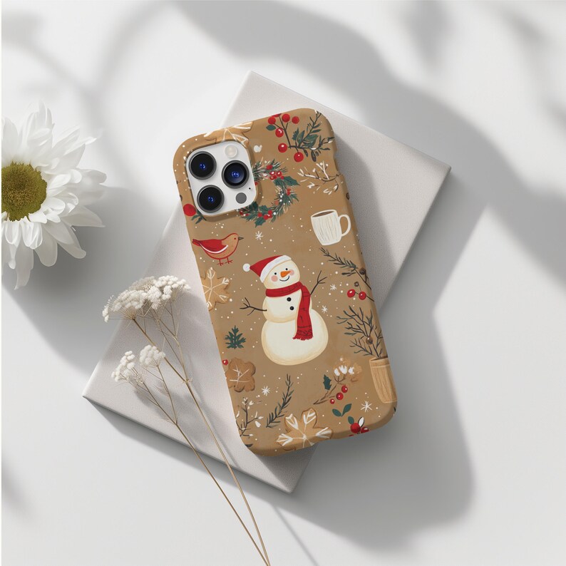 May include: A tan phone case featuring a festive Christmas design. The case is adorned with illustrations of a snowman wearing a red scarf and hat, a red bird, gingerbread cookies, and sprigs of holly. A white mug and a wreath are also featured.