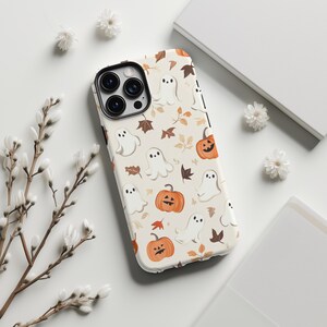May include: A phone case with a Halloween theme. The case features a pattern of white ghosts, orange pumpkins, and brown leaves on a cream background. The case has a black border and a camera cutout.