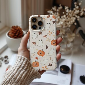 May include: A beige phone case featuring a repeating pattern of white ghosts, orange pumpkins, and brown autumn leaves. The case has a glossy finish and is designed to fit a smartphone. The case is held by a person.