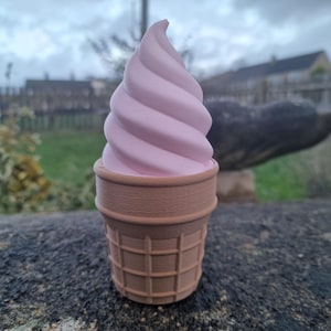 May include: A pink soft-serve ice cream cone with a waffle cone base. The ice cream has a swirled design. The cone is light brown. The background is blurred, showing a cloudy sky and a residential area.