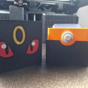 May include: Two black rectangular boxes. One has a gold ring and red eye shapes. The other has an orange interior and a white circular element. Both are likely 3D-printed items.