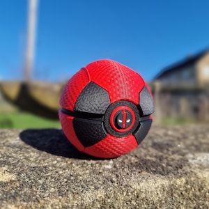 May include: A red and black ball with a Deadpool logo. The ball has a textured surface and is made of multiple panels. The Deadpool logo is in the center of the ball.