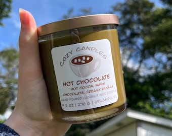 Könnte beinhalten: Eine Kerze im Glas mit kupferfarbenem Metalldeckel und weißem Etikett, das eine Tasse heiße Schokolade mit Marshmallows zeigt. Das Etikett trägt die Aufschrift COZY CANDLES, HOT CHOCOLATE, HOT COCOA, WARM CHOCOLATE, CREAMY VANILLA, HAND POURED COCONUT WAX CANDLE, 9.5 OZ | 270 G | ORLANDO, FL.