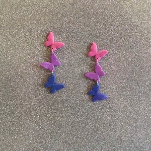 May include: A pair of dangle earrings featuring three layered butterflies. The top butterfly is pink, the middle butterfly is purple, and the bottom butterfly is blue.