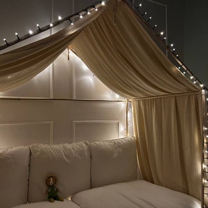 Puede incluir: Una cama con dosel blanco con una cubierta de tela beige y luces de cadena. La cama tiene una cabecera blanca y un edred&oacute;n blanco. Un peque&ntilde;o animal de peluche est&aacute; sentado en la cama.
