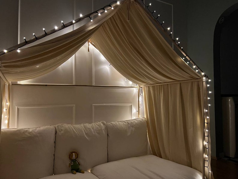 Puede incluir: Una cama con dosel blanco con una cubierta de tela beige y luces de cadena. La cama tiene una cabecera blanca y un edred&oacute;n blanco. Un peque&ntilde;o animal de peluche est&aacute; sentado en la cama.