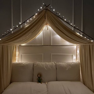 Puede incluir: Una cama con dosel blanco con una cubierta de tela beige y luces de cadena. La cama tiene una cabecera y un piecero blancos y un colchón blanco. Hay un animal de peluche en la cama.