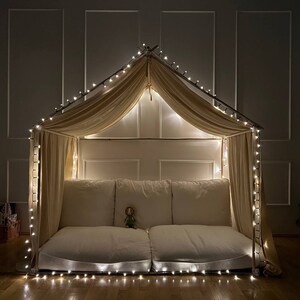 Puede incluir: Un acogedor fuerte interior hecho con una cubierta de tela beige y luces de cadena. El fuerte est&aacute; instalado en un suelo de madera y tiene un colch&oacute;n blanco mullido en el interior.