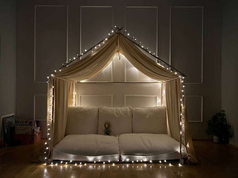 Puede incluir: Un acogedor fuerte interior hecho con una cubierta de tela beige y luces de cadena. El fuerte est&aacute; instalado en un suelo de madera y tiene un colch&oacute;n blanco mullido en el interior.