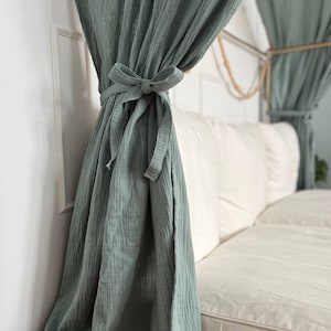 Pu&ograve; includere: Una tenda in cotone verde salvia con un fiocco annodato in cima, appesa davanti a un divano bianco.