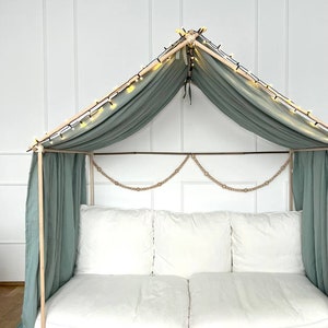 Pu&ograve; includere: Un divano bianco con due cuscini &egrave; all'interno di una tenda di stoffa verde chiaro con luci a catena. La tenda &egrave; fatta di pali di legno e ha una ghirlanda di corda appesa in cima.