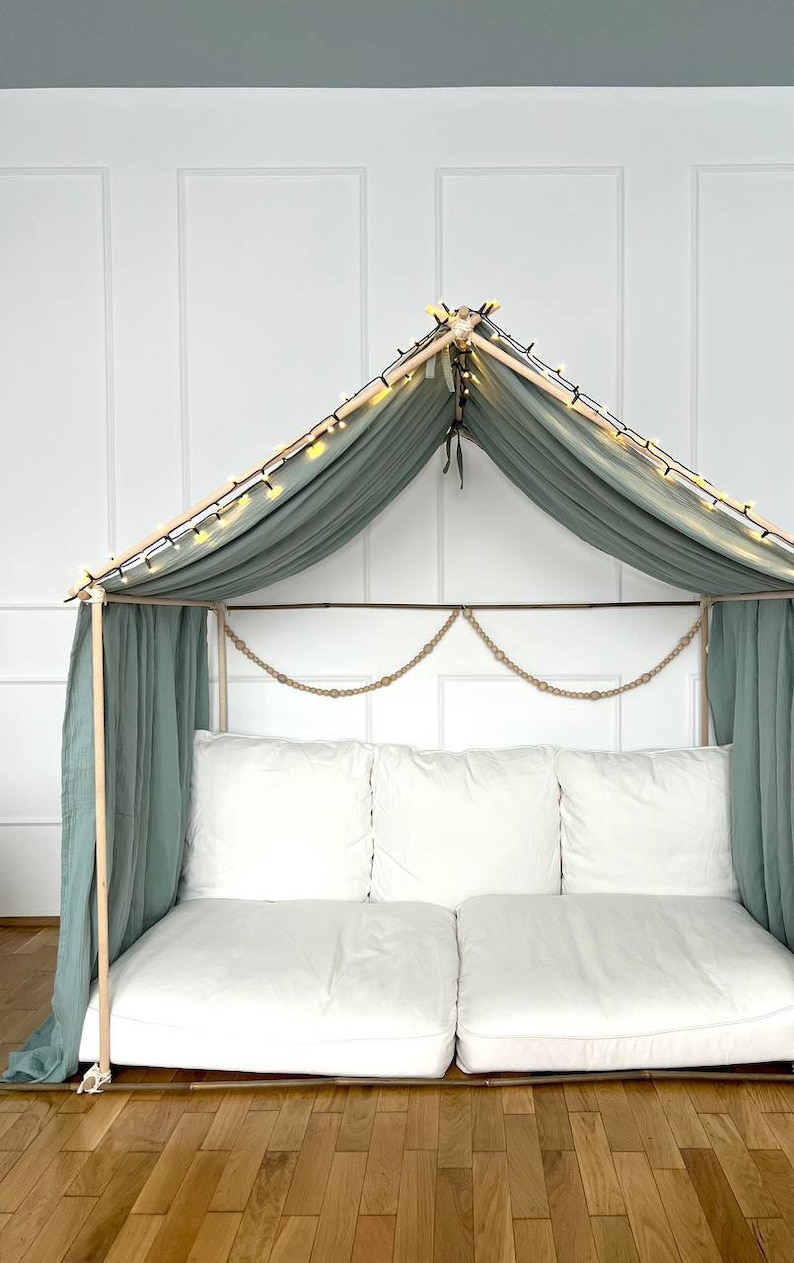 Pu&ograve; includere: Un divano bianco con due cuscini &egrave; all'interno di una tenda di stoffa verde chiaro con luci a catena. La tenda &egrave; fatta di pali di legno e ha una ghirlanda di corda appesa in cima.