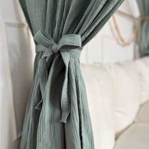 Pu&ograve; includere: Una tenda in cotone verde salvia con un fiocco annodato in basso. La tenda &egrave; appesa a un'asta e copre parzialmente un divano bianco.