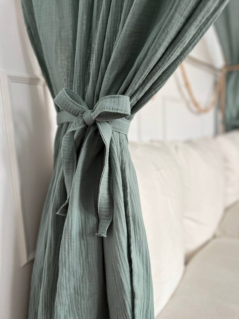 Pu&ograve; includere: Una tenda in cotone verde salvia con un fiocco annodato in basso. La tenda &egrave; appesa a un'asta e copre parzialmente un divano bianco.