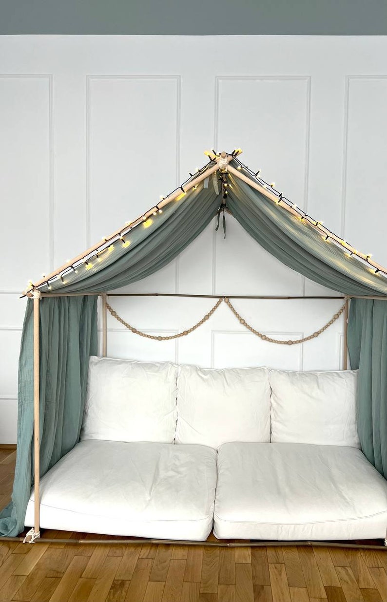 Pu&ograve; includere: Un baldacchino in tessuto verde chiaro drappeggiato su una struttura in legno con luci a catena. La struttura &egrave; fissata a un divano bianco con due cuscini bianchi. Il divano &egrave; su un pavimento in legno.