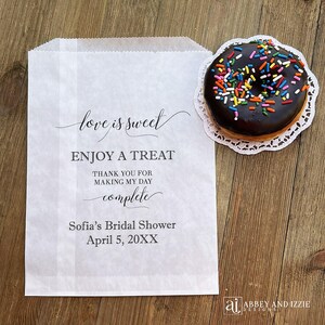 Puede incluir: Una bolsa de papel blanca con el texto "love is sweet" y "ENJOY A TREAT". Una rosquilla glaseada con chocolate y chispas de colores descansa sobre un encaje de papel blanco. La bolsa est&aacute; personalizada para una despedida de soltera.