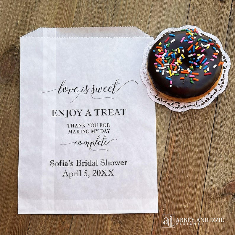 Puede incluir: Una bolsa de papel blanca con el texto "love is sweet" y "ENJOY A TREAT". Una rosquilla glaseada con chocolate y chispas de colores descansa sobre un encaje de papel blanco. La bolsa est&aacute; personalizada para una despedida de soltera.