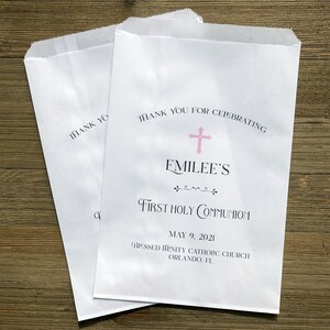May include: Two white paper treat bags. One bag reads "Thank You" and the other reads "Thank You For Celebrating" above "Emilee's First Holy Communion" with a pink cross. The date May 9, 2021 is printed on the bag, along with the church name.
