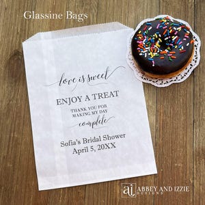 Puede incluir: Bolsa de papel glassine blanca con el texto "love is sweet" y "Enjoy a treat". Una rosquilla glaseada con chocolate y chispas de colores est&aacute; sobre un mantelito blanco junto a la bolsa. La bolsa es para la despedida de soltera de Sof&iacute;a.