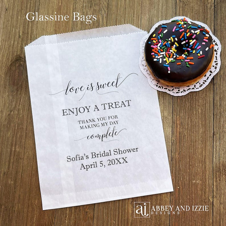 Puede incluir: Bolsa de papel glassine blanca con el texto "love is sweet" y "Enjoy a treat". Una rosquilla glaseada con chocolate y chispas de colores est&aacute; sobre un mantelito blanco junto a la bolsa. La bolsa es para la despedida de soltera de Sof&iacute;a.