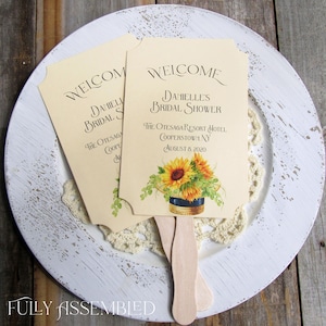 May include: Two yellow and cream-colored paper fans with wooden handles. The fans are decorated with sunflowers and the text "Welcome Danielle's Bridal Shower The Otesaga Resort Hotel Cooperstown, NY August 8, 2020".