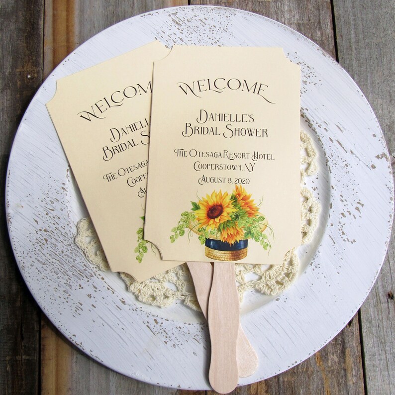 May include: Two yellow and white paper fans with wooden handles. The fans have a sunflower design and text that reads "Welcome Danielle's Bridal Shower The Otesaga Resort Hotel Cooperstown, NY August 8, 2020".