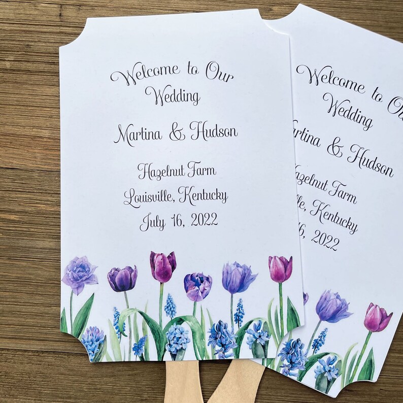 Op de afbeelding: Twee witte bruiloftsvaandels met paarse en blauwe bloemenmotieven. De tekst op de vaandels luidt "Welcome to Our Wedding Martina & Hudson Hazelnut Farm Louisville, Kentucky July 16, 2022".