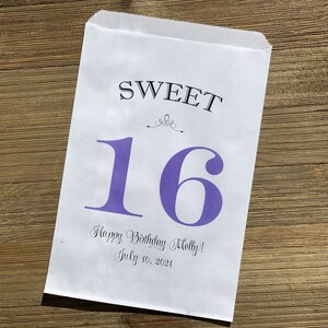 Op de afbeelding: Een witte papieren cadeauzak met de woorden "SWEET 16" in zwart en paars gedrukt. De zak bevat ook de tekst "Happy Birthday Molly! July 10, 2021" in een scriptlettertype. De zak ligt op een houten oppervlak.