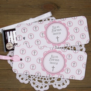 May include: Two pink and white favor tags with a cross pattern and a pink circle with the text "Ella's Baptism 08.07.21".
