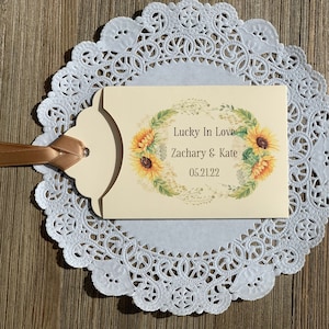 Puede incluir: Una tarjeta de color crema con un dise&ntilde;o de corona de girasoles. El texto dice "Lucky In Love Zachary & Kate 05.21.22".