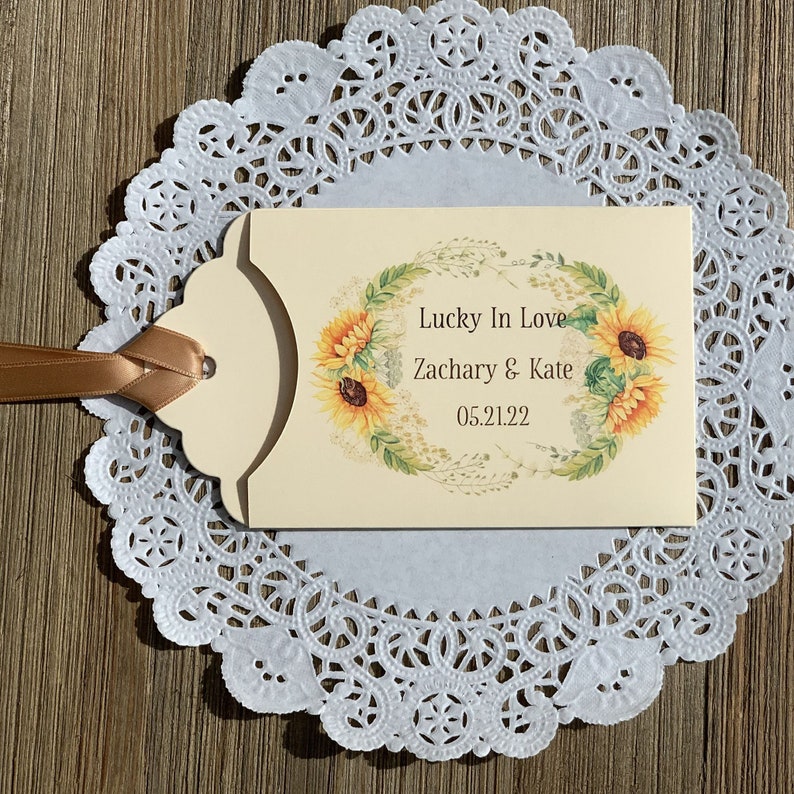 Puede incluir: Una tarjeta de color crema con un dise&ntilde;o de corona de girasoles. El texto dice "Lucky In Love Zachary & Kate 05.21.22".