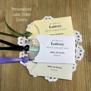 May include: Two white cards with the text "If life is a lottery, we've already won" and the names "Abbey & Justin" and the date "06-29-24". The cards are decorated with a green and purple ribbon and a small lottery ticket.