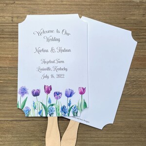 Op de afbeelding: Een witte bruiloftswaaier met een bloemenontwerp met paarse en blauwe tulpen. De waaier heeft de tekst "Welcome to Our Wedding Martina & Hudson Hazelnut Farm Louisville, Kentucky July 16, 2022" erop gedrukt.