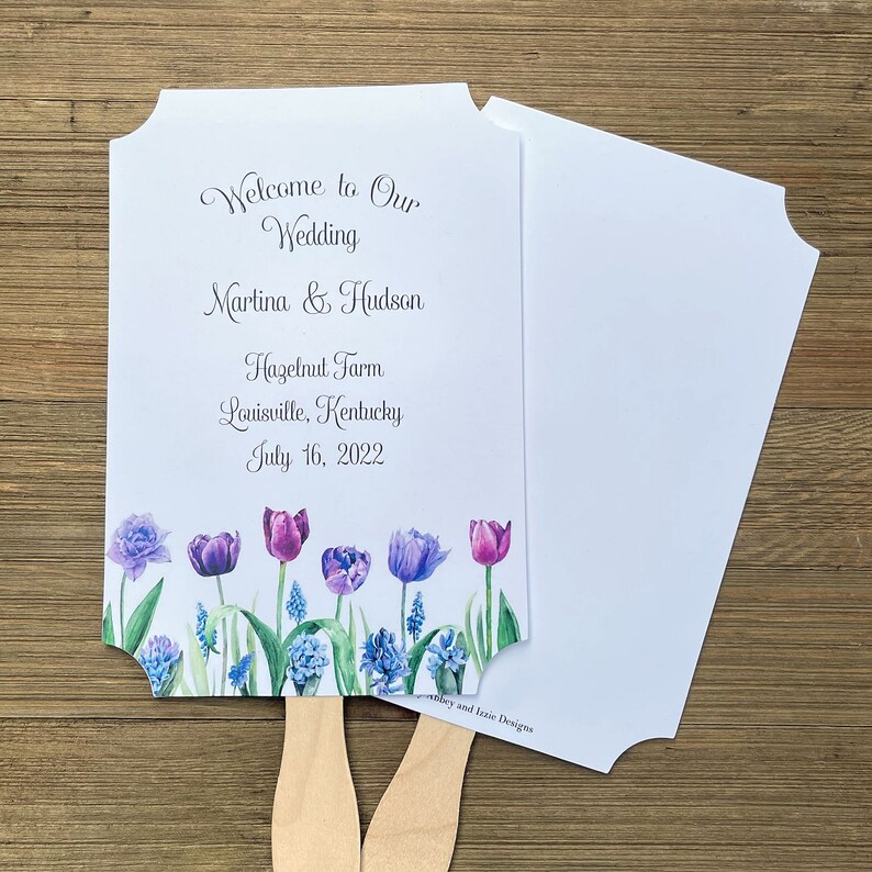 Op de afbeelding: Een witte bruiloftswaaier met een bloemenontwerp met paarse en blauwe tulpen. De waaier heeft de tekst "Welcome to Our Wedding Martina & Hudson Hazelnut Farm Louisville, Kentucky July 16, 2022" erop gedrukt.