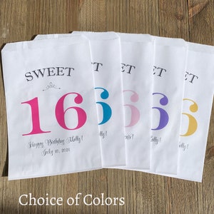 Op de afbeelding: Witte papieren traktatiezakken met "SWEET 16" in verschillende kleuren bedrukt. De zakken bevatten de tekst "Happy Birthday Molly! July 10, 2021" en "Choice of Colors". De zakken liggen op een houten oppervlak.