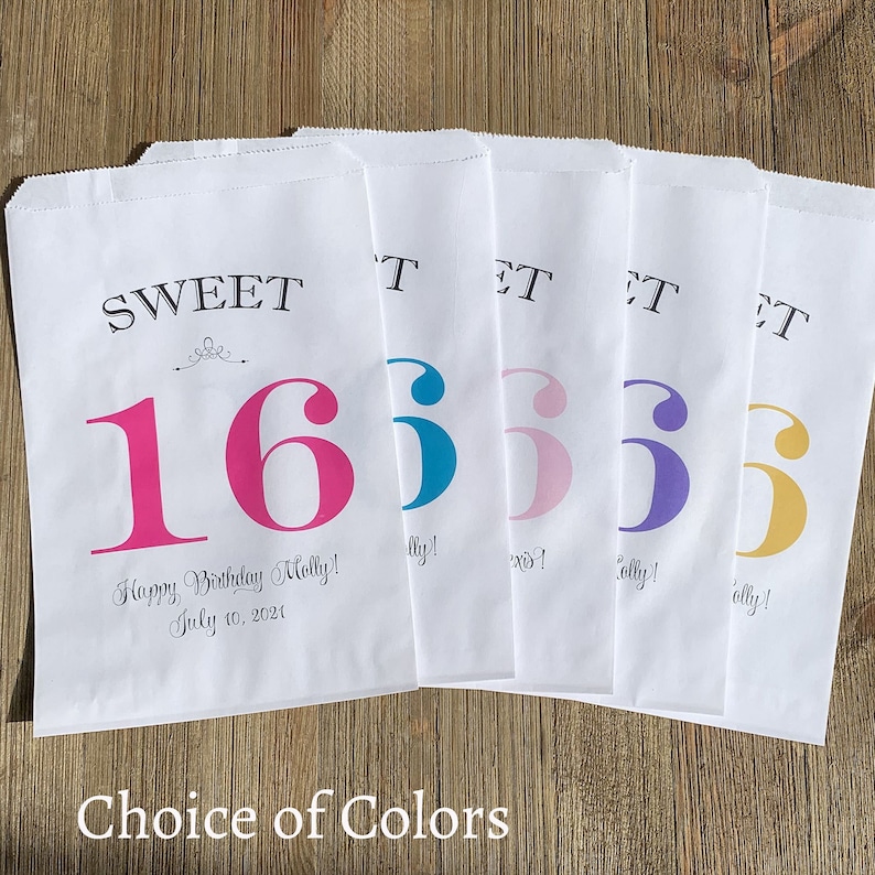 Op de afbeelding: Witte papieren traktatiezakken met "SWEET 16" in verschillende kleuren bedrukt. De zakken bevatten de tekst "Happy Birthday Molly! July 10, 2021" en "Choice of Colors". De zakken liggen op een houten oppervlak.