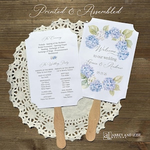 May include: Wedding fan featuring a floral design with blue hydrangeas and the text "Welcome to our wedding Grace & Andrew". The fan includes the wedding date and details of the ceremony and wedding party. The fan is made of paper and wood.