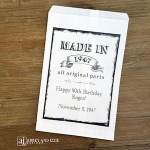 K&ouml;nnte beinhalten: Eine wei&szlig;e Papiert&uuml;te mit schwarzem Rand und Text, der "MADE IN 1947" und "Happy 80th Birthday Roger!" auf einer Holzoberfl&auml;che zeigt. Die T&uuml;te enth&auml;lt auch den Text "all original parts" und "November 5, 1947."