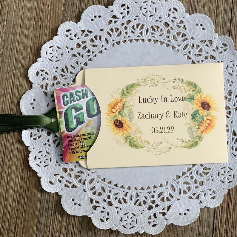 Puede incluir: Un sobre amarillo con un dise&ntilde;o de corona de girasoles. El sobre tiene el texto "Lucky In Love Zachary & Kate 05.21.22". Un peque&ntilde;o boleto de raspar colorido est&aacute; unido al sobre con una cinta verde.