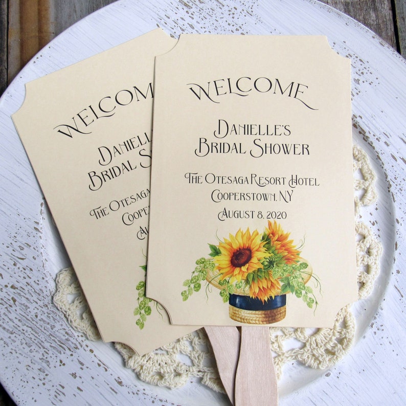 May include: Two cream-colored paper fans with scalloped edges. The top fan has the text "WELCOME DANIELLE'S BRIDAL SHOWER THE OTESAGA RESORT HOTEL COOPERSTOWN, NY AUGUST 8, 2020" and a sunflower illustration. The bottom fan has the text "WELCOME DANIELLE BRIDAL SHOWER THE OTESAGA COOPER" and a green leaf illustration.