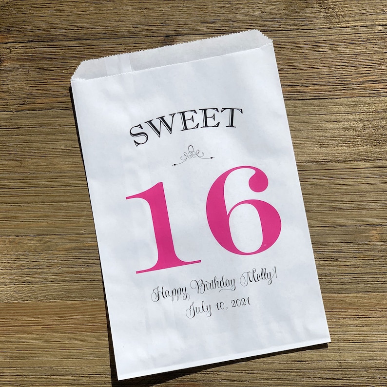 Op de afbeelding: Witte papieren traktatiezak met de woorden "SWEET 16" in zwart en roze. De zak bevat ook de tekst "Happy Birthday Molly! July 10, 2021" in een scriptlettertype. De zak ligt op een houten oppervlak.