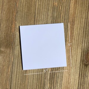 Może przedstawiać: Biała, pusta karta o wymiarach 12,7 cm x 12,7 cm. Karta leży płasko na drewnianej powierzchni.