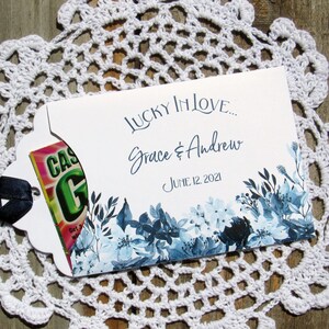 May include: A white wedding favor envelope with a blue floral design. The envelope has a blue ribbon and a small, colorful card that says "Lucky in Love... Grace & Andrew June 12, 2021".
