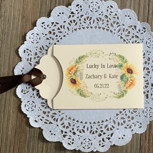 Puede incluir: Una tarjeta de color crema con un dise&ntilde;o de corona de girasoles. El texto dice "Lucky in Love Zachary & Kate 05.21.22". La tarjeta est&aacute; unida a una cinta marr&oacute;n.