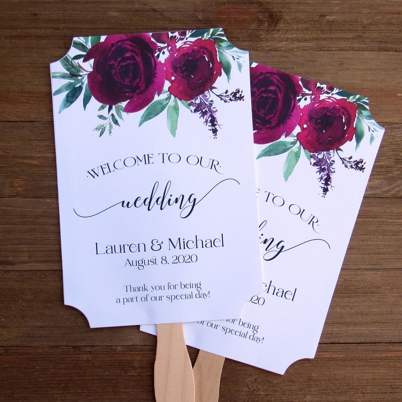 May include: Two white wedding fans with burgundy floral designs. The text on the fans reads "Welcome to our wedding", "Lauren & Michael", "August 8, 2020", and "Thank you for being a part of our special day!"