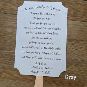 May include: A light blue wedding fan with a message to family and friends. The message reads "To Our Family & Friends It means the world to us to have you here. Thank you for your support, encouragement and love and laughter you have contributed to our lives. You are our teachers, partners in crime, guides and favorite people in the whole world. We hope you enjoy today's celebration and those we'll share for years to come. with love, Kristen & Jack August 13, 2022". The fan is on a wooden surface with the word "Gray" written below it.